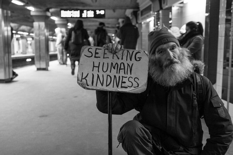 Bearded old man sitting in subway station holds sign that says, "Seeking Human Kindness."