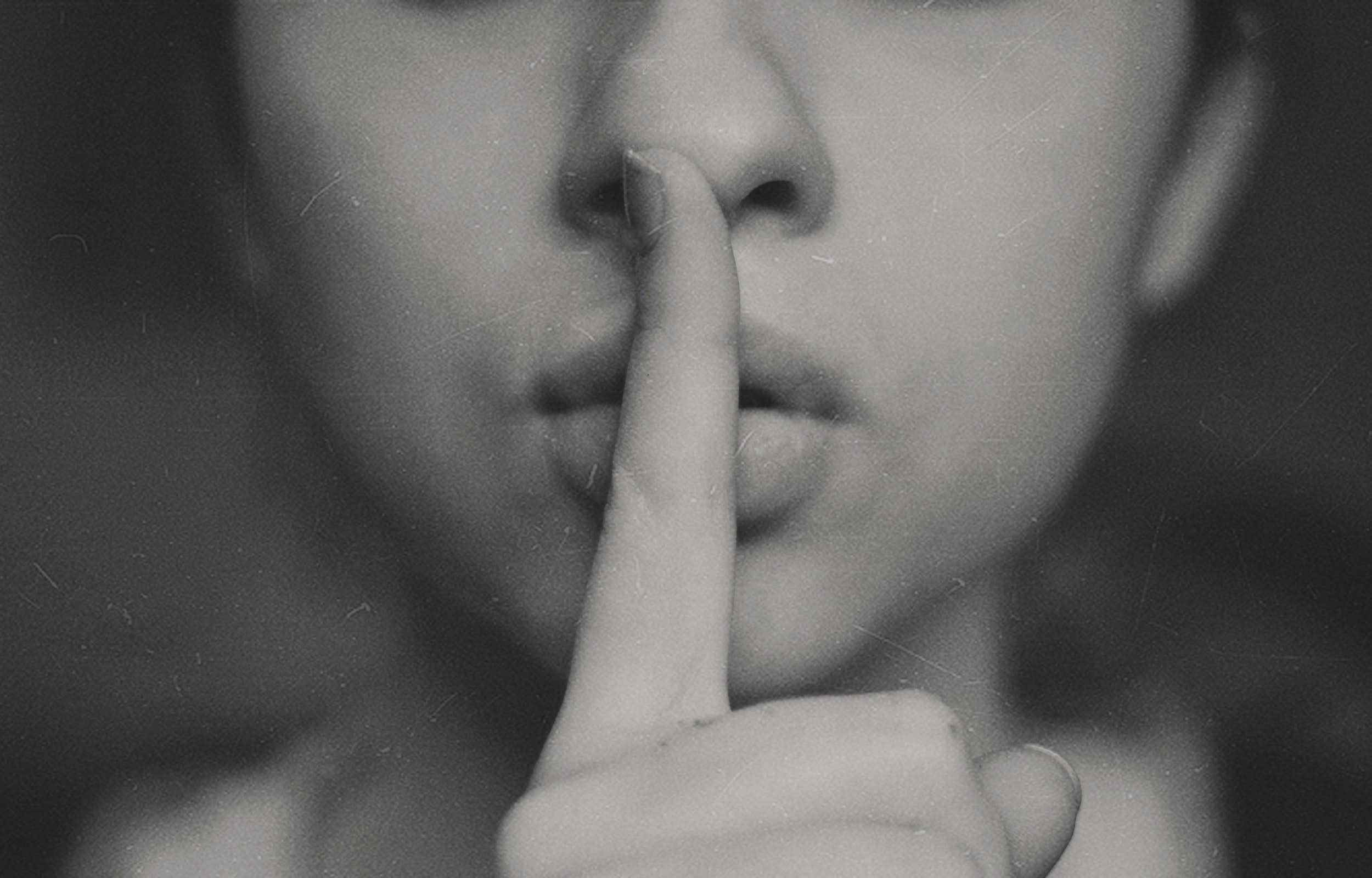 Close up of lower half of person's face with their index finger in front of their mouth in a "ssh" gesture.